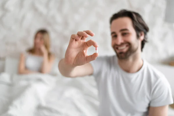 Cheerful young guy holding pill for erectile dysfunction, smiling wife sitting in bed on background, selective focus — Fotografie, imagine de stoc