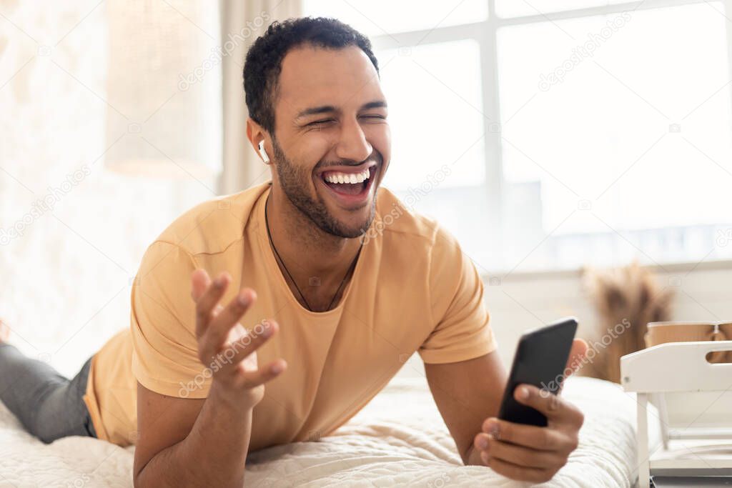 Arabic Guy Listening Music Wearing Earbuds Holding Smartphone In Bedroom
