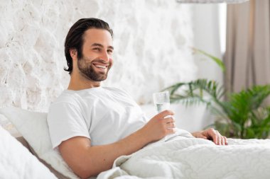 Happy young man taking glass of water in bed
