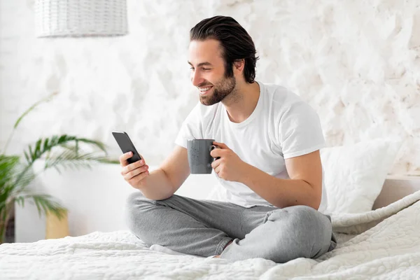 Well-rested handsome guy chilling on bed at home, using smartphone — Stock Photo, Image