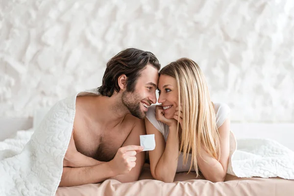 Safe sex. Young spouses holding condom, lying on bed under blanket, looking at each other with love at home, copy space — Stockfoto