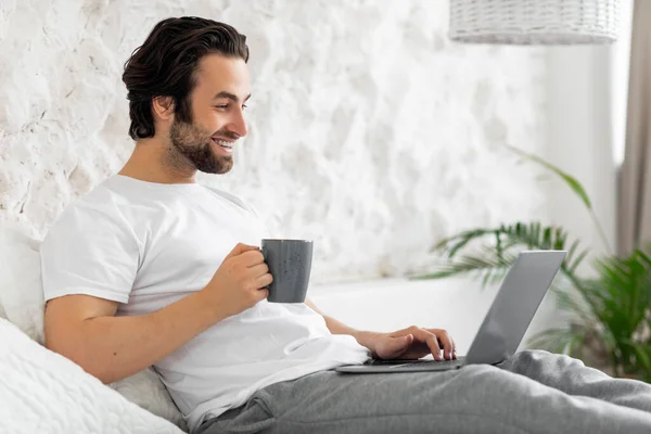 Handsome young guy using laptop in bed, drinking coffee — Zdjęcie stockowe