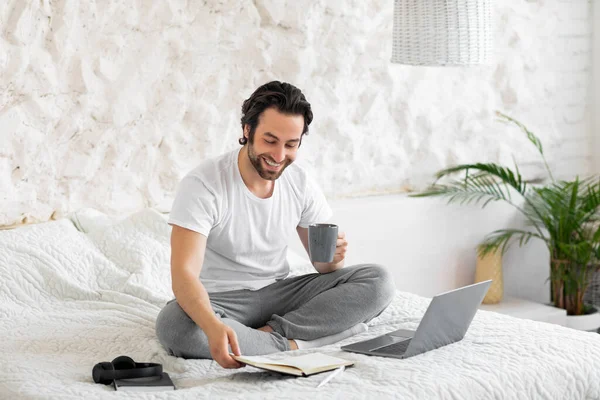 Relaxed young man checking his agenda, drinking coffee in bed — Foto de Stock