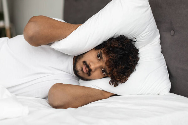 Irritated eastern man laying in bed, covering head with pillow