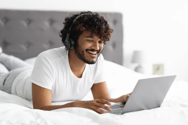 Relaxed eastern guy enjoying weekend in bed, using laptop — Foto Stock