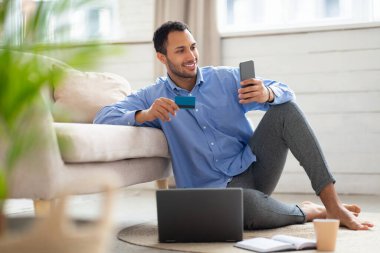 Evde cep telefonu ve kredi kartı kullanan gülümseyen bir Arap.