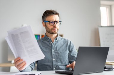 Focused mature man reading documents, having online business conference at office, sitting at workplace with laptop