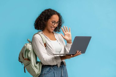 Sırt çantalı siyah bir kız öğrenci dizüstü bilgisayara el sallıyor, üniversite öğretmeniyle konuşuyor, görüntülü konuşma yapıyor ya da internet dersi veriyor.