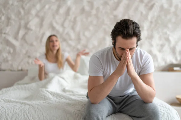 Young man feeling depressed, sitting on bed at home, his angry wife shouting at him, free space. Family problems concept — Stockfoto