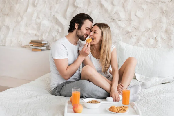 Piena lunghezza di bel ragazzo che alimenta croissant fresco alla sua ragazza, seduto sul letto a casa — Foto Stock