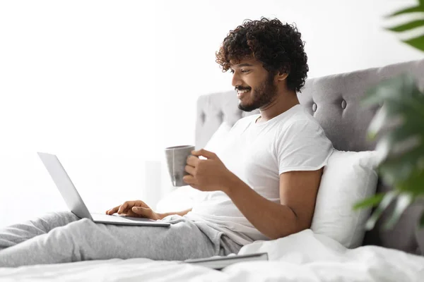 Entspannter indischer Mann trinkt Kaffee im Bett mit Laptop — Stockfoto