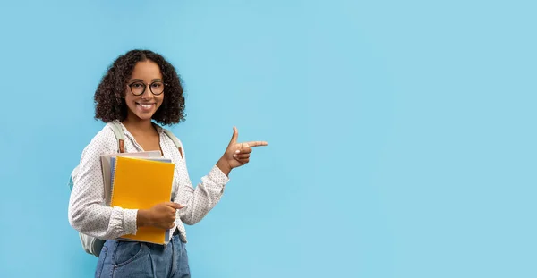 Beautiful black female student with backpack and notebooks pointing aside at empty space for your educational ad — Stockfoto