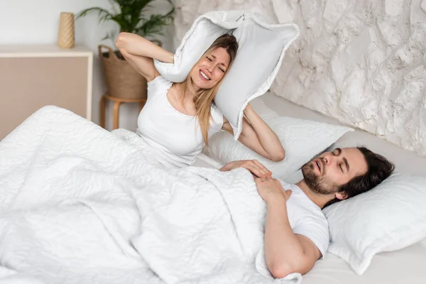 Snoring young guy and his frustrated girlfriend covering ears with pillow in bed at home —  Fotos de Stock