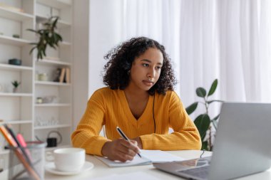 Focused young black woman in headset having online lesson on laptop, writing in notebook at home