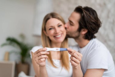 Happy young couple showing positive pregnancy test and kissing at home, selective focus clipart