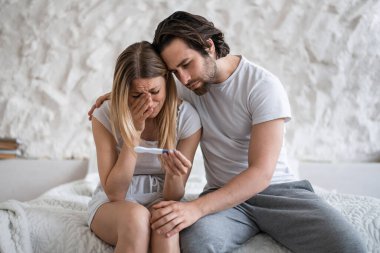 Upset young woman with negative pregnancy test crying on bed, loving husband supporting her at home