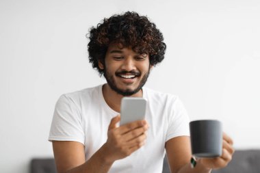 Closeup of curly eastern guy drinking tea, using phone