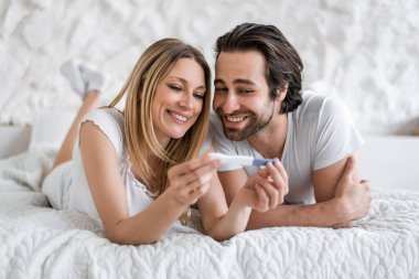 Mutlu genç kadın ve kocası evde pozitif hamilelik testiyle yatakta yatıyorlar.