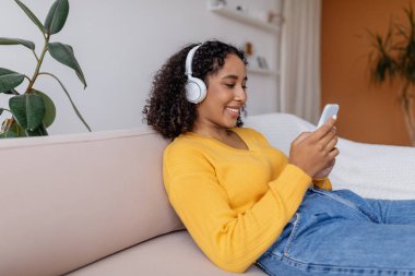 Kulaklık takmış, kanepede dinlenen, müzik dinleyen ya da akıllı telefondan film izleyen güzel siyahi bir kadın.