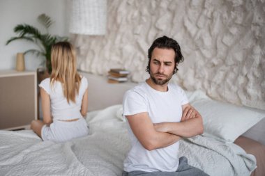 Unhappy young couple having relationship problems, sitting on opposite sides of bed, not looking at each other at home
