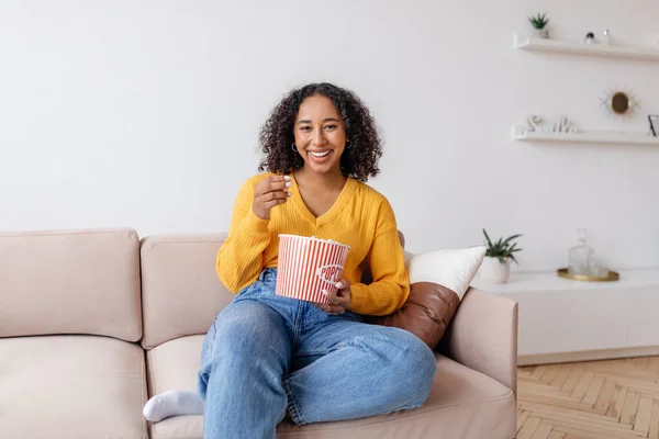 Cheery young black woman eating popcorn while watching TV on couch in living room —  Fotos de Stock