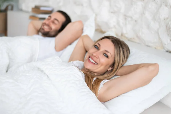 Happy young married couple relaxing on soft pillows in bed, enjoying lazy weekend morning at home, copy space — 스톡 사진