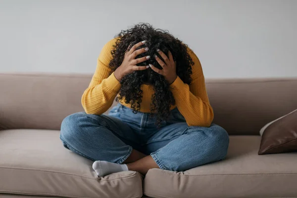Depressed young black woman sitting cross legged on sofa with head in her hands at home — Photo