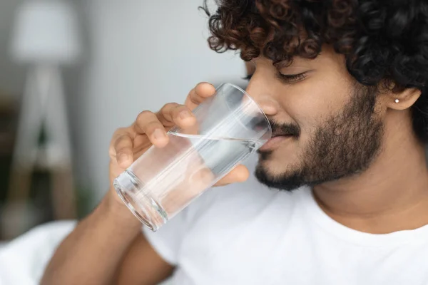Happy curly young man taking glass of water in bed — Stockfoto