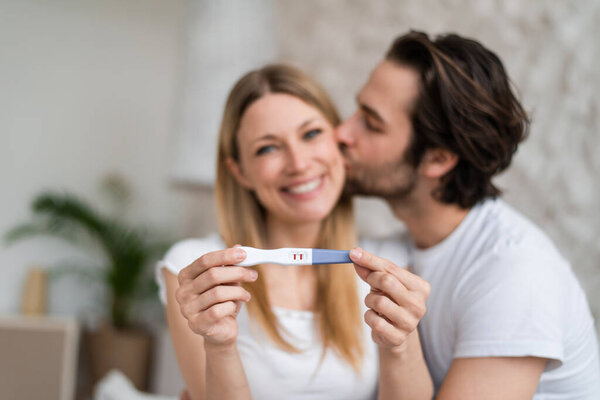 Happy young couple showing positive pregnancy test and kissing at home, selective focus