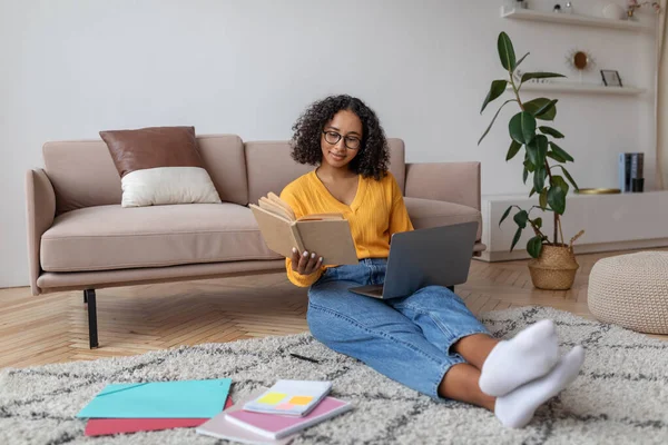 Happy young African American woman studying online from home, reading book, using laptop pc at home, free space — Zdjęcie stockowe