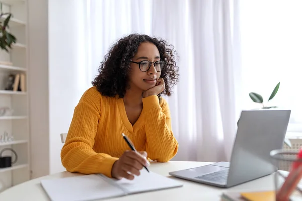 Positive young black female student having online lesson on laptop computer, writing down information at home — Stockfoto