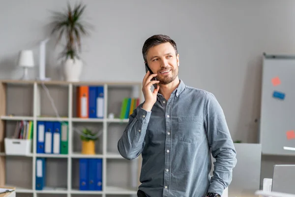 Positive mature man in office talking on mobile phone near workplace, having conversation with client — Foto de Stock