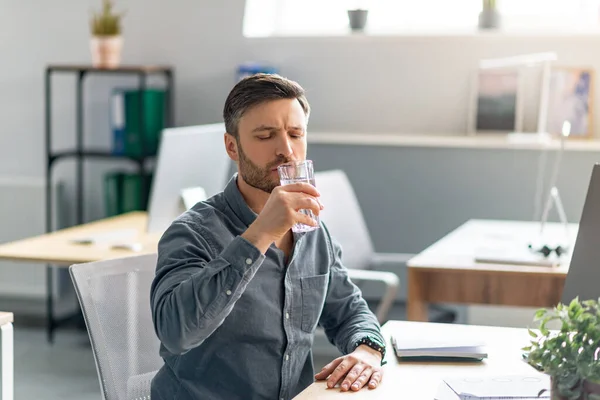 Mature male manager holding glass of water, drinking water while working on computer in office, copy space — ストック写真