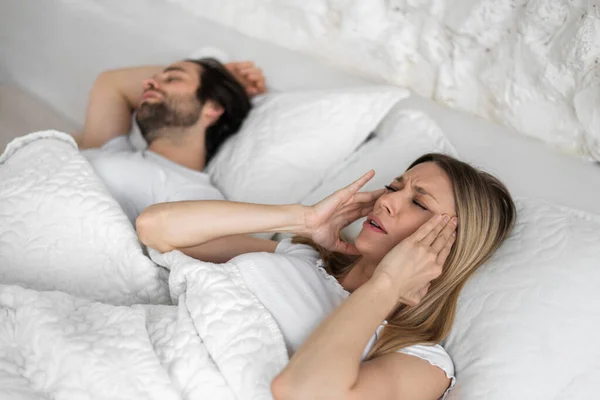興奮した若い女性に悩まされている夫がいびきをかく、家のベッドで耳を覆い、スペースをコピー — ストック写真