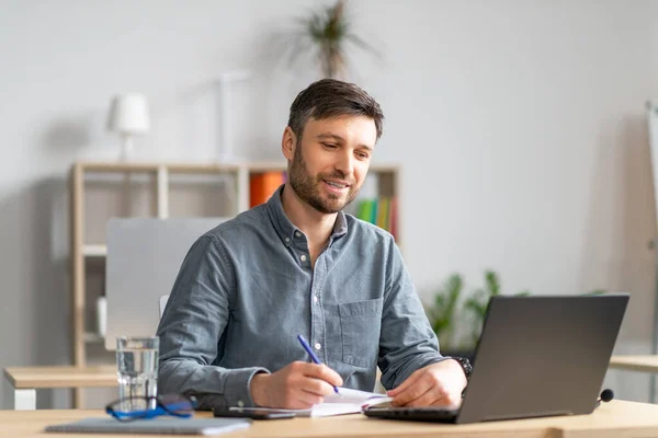 Happy mature entrepreneur man taking notes and using laptop, sitting at his workplace in office, copy space — стоковое фото