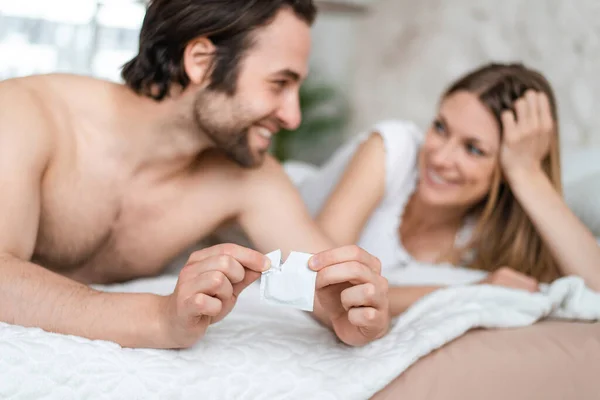 Joyeux jeune couple couché au lit et ouvrant préservatif, foyer sélectif. Concept de contrôle de la grossesse et des MTS — Photo