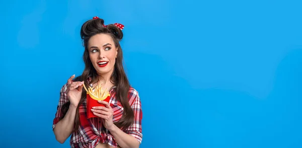 Charmig ung pinup kvinna i retro kläder äter pommes frites från förpackningen, titta på kopia utrymme på blå bakgrund — Stockfoto
