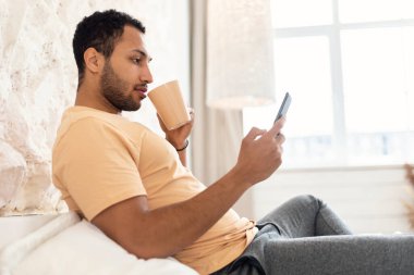 Orta Doğulu adam yatak odasında telefon kullanıyor ve kahve içiyor.