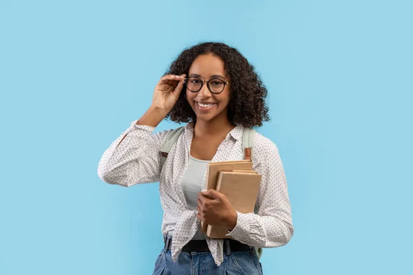 Портрет молодої чорношкірої студентки з книгами, які торкаються окулярів і посміхаються на камеру над синім студійним фоном — стокове фото