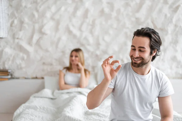 Young man holding pill, solving sexual dysfunction problems, happy woman sitting in bed and waiting for her husband — Stockfoto