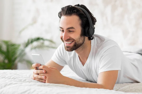 Happy guy using smartphone and headset, bedroom interior — Foto de Stock
