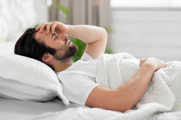 Upset young man waking up with headache, bedroom interior — Stock fotografie