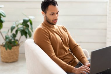 Arap adam evdeki koltukta dizüstü bilgisayar kullanıyor.