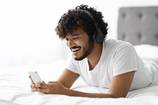 Joyful indian guy laying in bed with smartphone and headset — Foto Stock