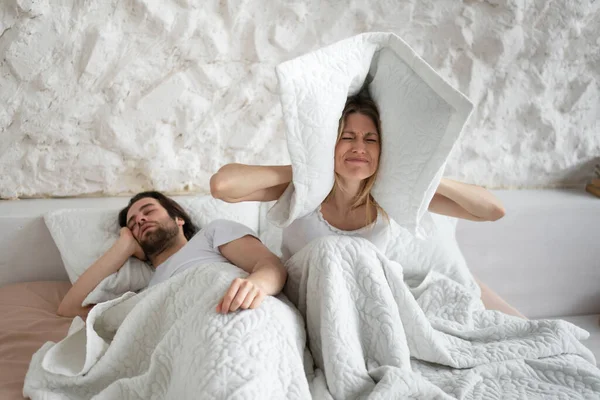Απογοητευμένη νεαρή γυναίκα καλύπτει τα αυτιά με μαξιλάρι, ενώ ο σύζυγός της ροχαλίζει στο κρεβάτι στο σπίτι, αντίγραφο χώρο — Φωτογραφία Αρχείου