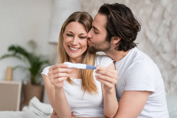 Iubirea cuplului tânăr se simte fericit cu privire la testul de sarcină pozitiv, sărutându-se și îmbrățișându-se acasă — Fotografie, imagine de stoc