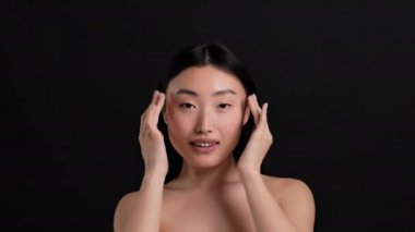 Beauty care habits. Young beautiful korean lady doing face lifting massage, touching smooth skin over black background