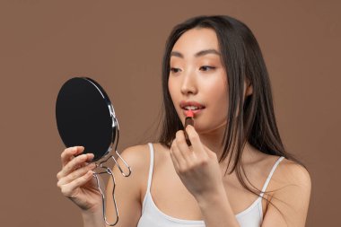 Genç Koreli kadın ayna ve ruj kullanıyor, çıplak makyaj yapıyor, kahverengi stüdyo arka planında duruyor.