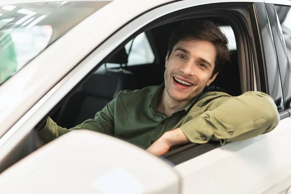 Happy millennial man sitting on drivers seat of new automobile after purchase, looking out window and smiling at dealership — Stock Fotó