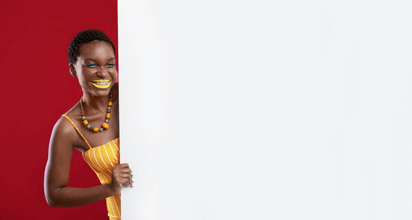 Cute short-haired young african american woman wearing stylish summmer look posing with horizontal white empty placard over red studio background, panorama with copy space, mockup
